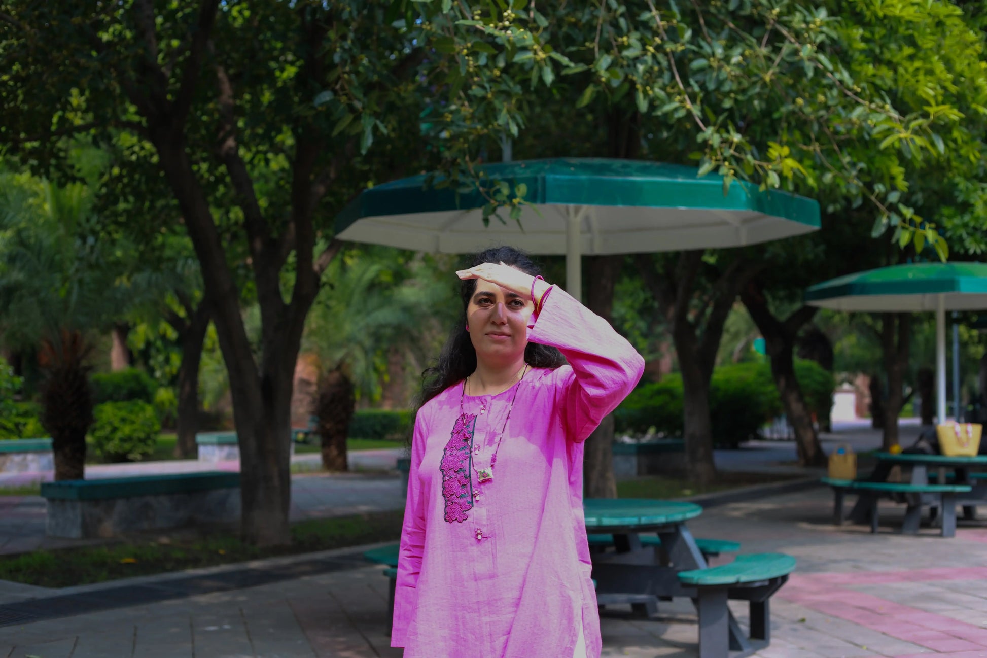 Woman in a pink traditional outfit standing in a park with green trees and benches.
